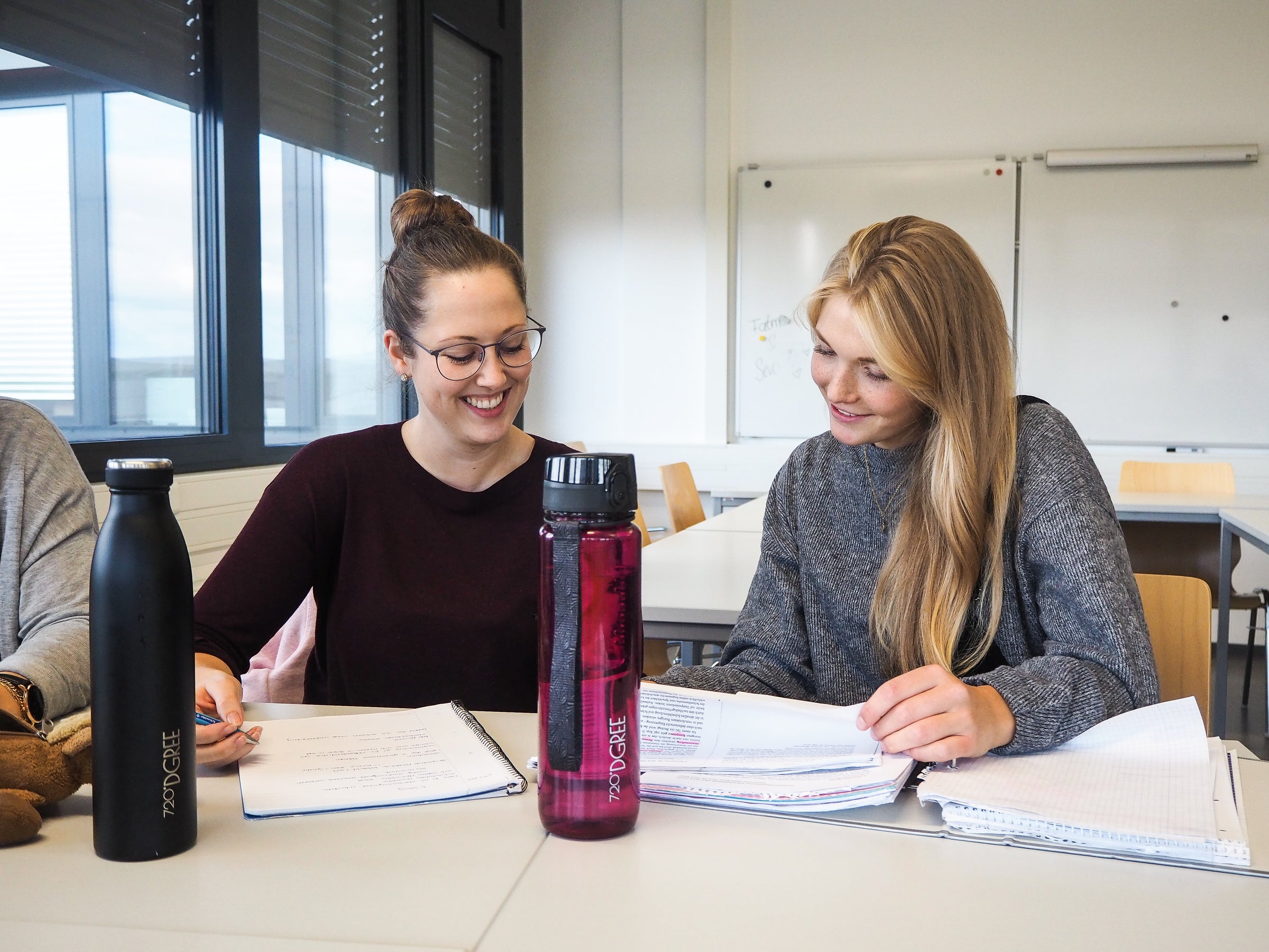 Zwei Studentinnen sitzen in einem Seminarraum nebeneinander und tauschen sich intensiv miteinander aus. Vor ihnen stehen rechts eine crystalClear uberBottle in Imperial Red und eine milkyBottle in Onyx Black. Die perfekte Unterstützung für Dein Studium.